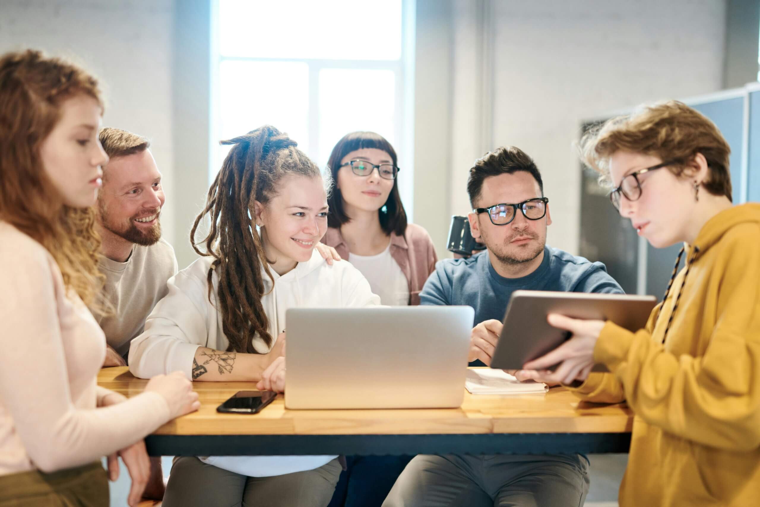 A diverse group of six people gather around a table with laptops and notepads, actively discussing.