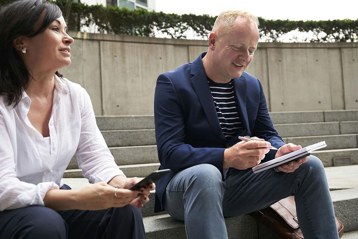 Coaching session between man and woman sitting in a courtyard. The woman is on her phone and the man is looking at his notebook. He's using the Autocratic type of coaching.