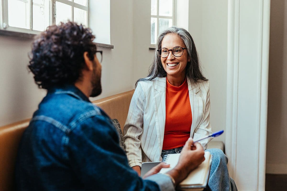 older woman employee sitting on a bench in a hallway with a male colleague during a mentoring engagement meeting