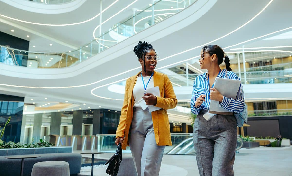 Two women walking in the lobby of a modern office talking during a mentoring meeting.