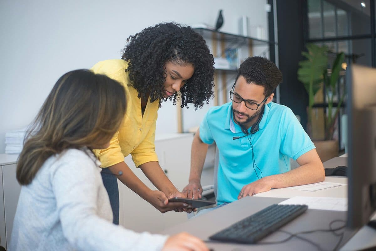 Two women employees and a male employee sit in an office looking at a tablet. They are reviewing the types of coaching offered in their coaching program in their organization.